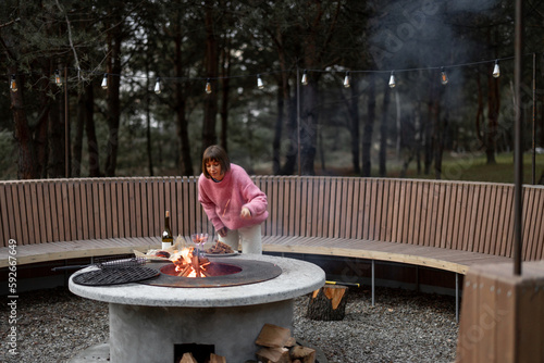 Young woman prepares food on beautiful outdoor bbq area with fire and round bench in pine forest at dusk. Luxury lifestyle at countryside concept