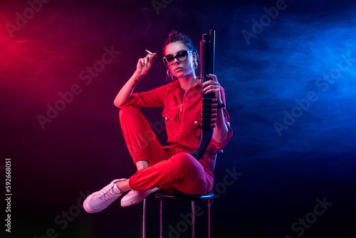 Fototapeta Naklejka Na Ścianę i Meble -  sexy bully girl smokes a cigarette with a shotgun on a dark background in neon light and a haze of copy paste