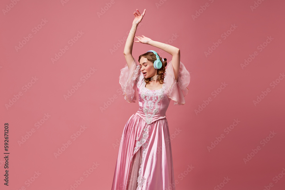 Naklejka premium Shot of adorable blond queen wearing headphones and dress listening music over pink studio background. Modern princess