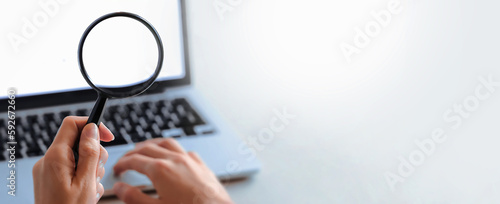 Hand holding magnifying glass in front of a laptop screen. Symbol of web browsing or zooming displeyed something on screen. Paying attention on detail.