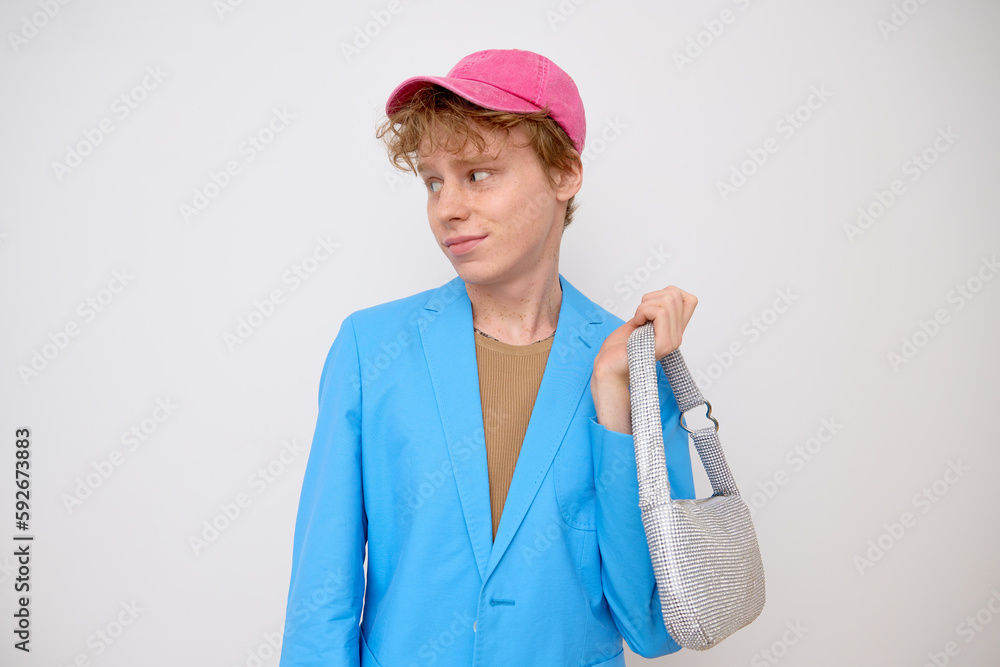 Young gay ginger curly haired man blogger wearing fashionable outfit with pink cap and blue blazer and carrying a silver sparkly handbag looking away smiling and posing for photo over white backdrop. Young gay ginger curly haired man blogger wearing fashionable outfit with pink cap and blue blazer and carrying a silver sparkly handbag looking away smiling and posing for photo over white backdrop.