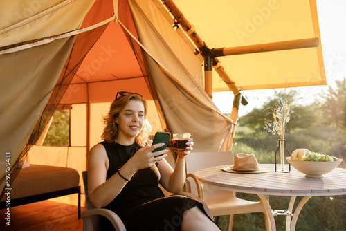 Happy Woman freelancer use phone and drinking cocktail call while relaxing on cozy glamping tent in sunny day. Luxury camping tent for outdoor summer holiday and vacation. Lifestyle concept