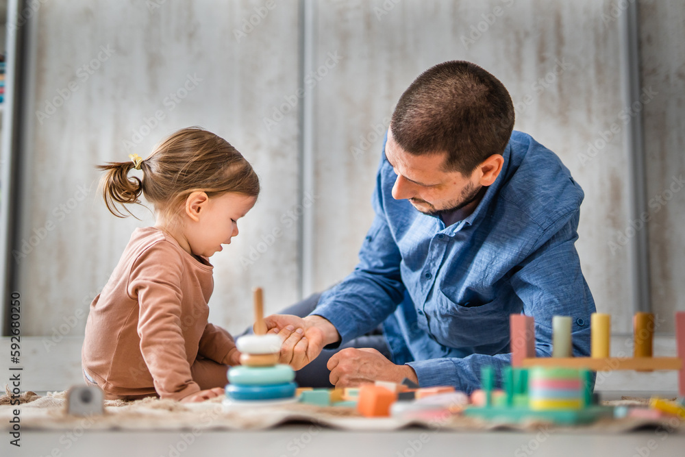 Naklejka premium One man caucasian playing at home with his daughter toddler on the floor with wooden toys, modern parenthood lifestyle single parent concept 