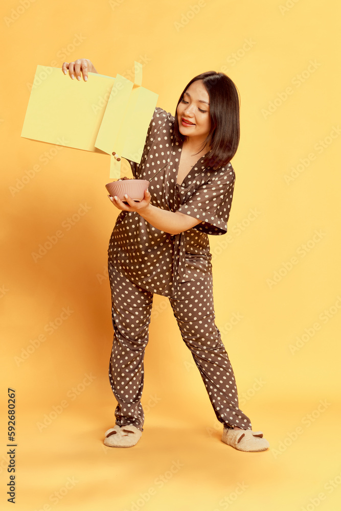 Full-length portrait of young, positive, asian girl in comfortable homewear, putting cereal into bowl against yellow studio background. Morning breakfast. Concept of emotions, lifestyle, youth Full-length portrait of young, positive, asian girl in comfortable homewear, putting cereal into bowl against yellow studio background. Morning breakfast. Concept of emotions, lifestyle, youth