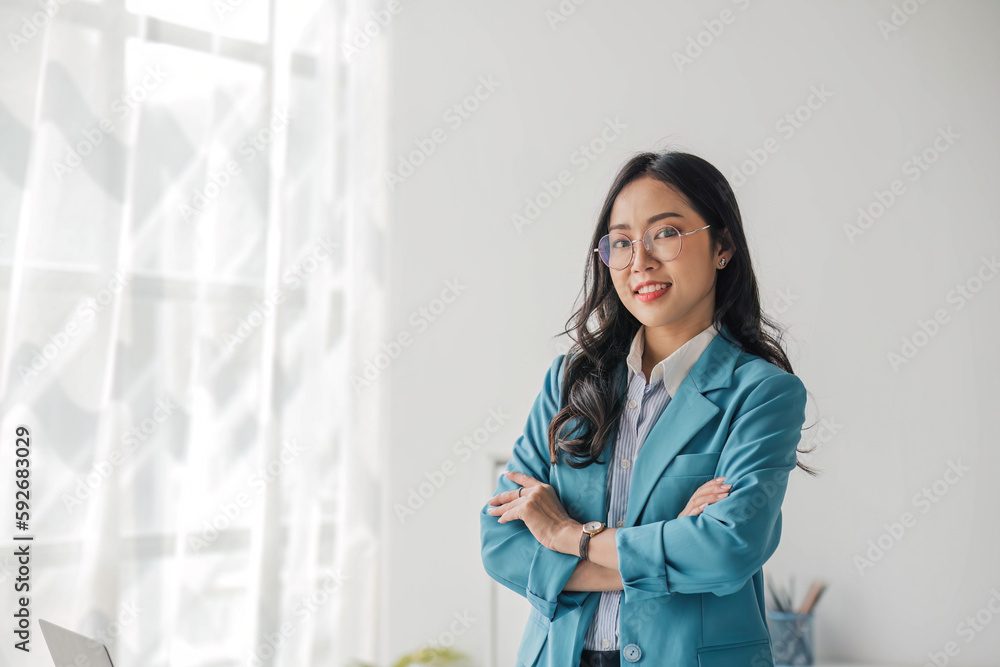 Portrait of young Asian woman in casual clothes standing with arms crossed, smiling and looking at camera while standing in modern office. Portrait of young Asian woman in casual clothes standing with arms crossed, smiling and looking at camera while standing in modern office.