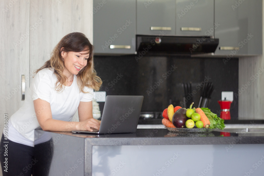 A beautiful woman smiles as she works on her laptop at a table in her kitchen at home A beautiful woman smiles as she works on her laptop at a table in her kitchen at home