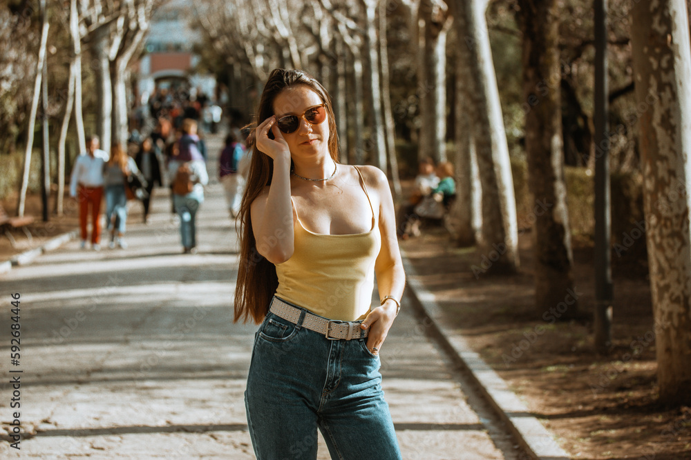 A beautiful European white brunette girl with long brown hair in jeans is standing outside in a city park in the summer. People in the street. A smiling young woman posing outdoors. Travel vacations.
