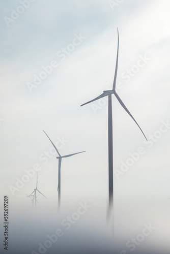 Windmills with operating rotor blades producing renewable energy in countryside. Wind turbines hidden in thick smog in cold early morning