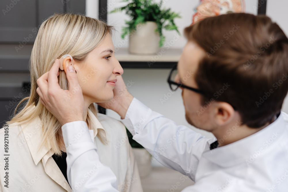 A young attractive otolaryngologist doctor gives a consultation to a female patient. A doctor explains how to wear a hearing aid to a woman - obrazy, fototapety, plakaty 