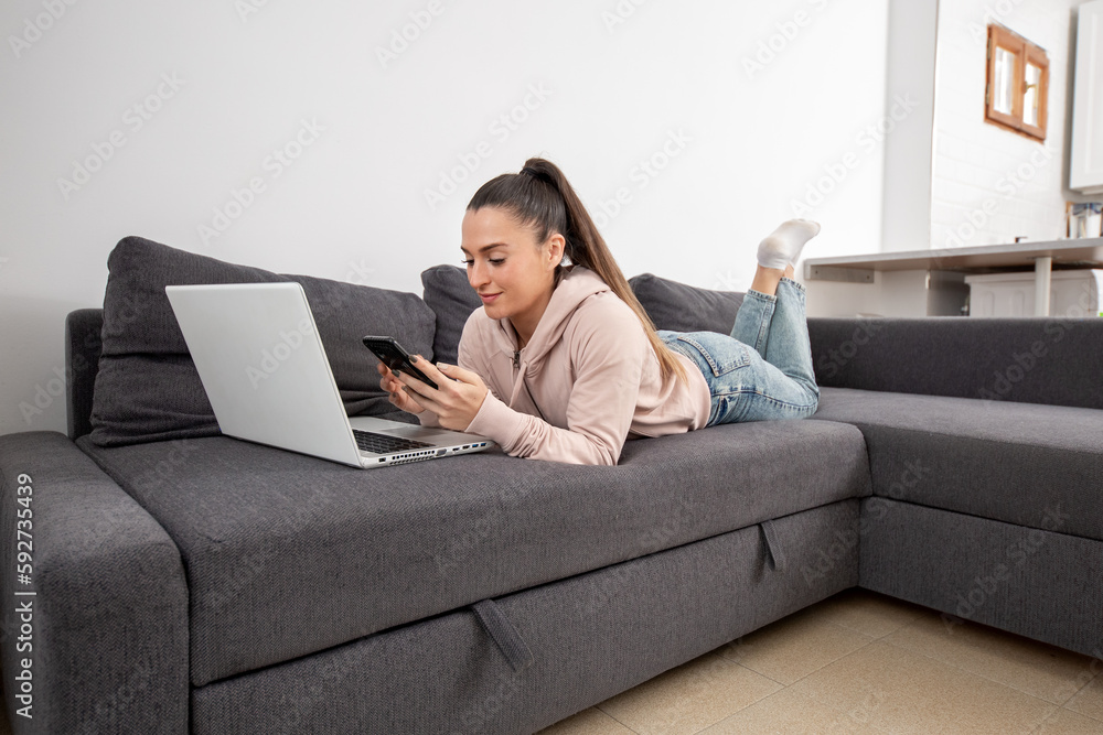Woman lying on the sofa looking at her smartphone in front of her laptop Woman lying on the sofa looking at her smartphone in front of her laptop