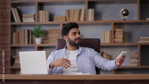 bearded Hindu man freelancer get warm working typing on laptop and turn on remote control changing temperature on air conditioner cooling system or humidifier at computer desk in the office