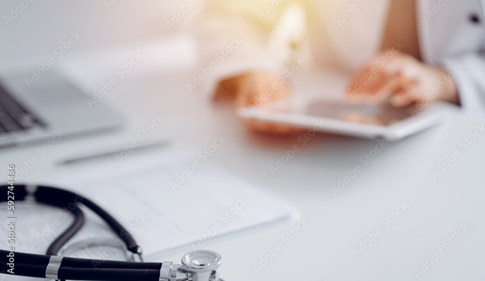 Stethoscope lying at doctor's working desk, close up. Physician uses tablet computer at the background. Medicine concept Stethoscope lying at doctor's working desk, close up. Physician uses tablet computer at the background. Medicine concept