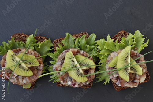 Three slices of rye bread with herring tartare, garnished with kiwi slices.