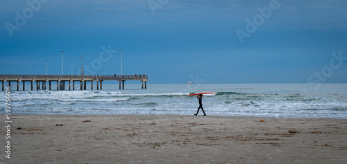 Obraz na płótnie PORT ARANSAS, TX - 9 FEB 2023: Young surfer carries a surfboard on the beach, near the water of the Gulf of Mexico and a pier, in evening light. w salonie
