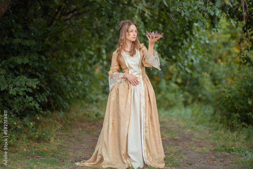 Beautiful young woman in a historical gold dress with a crown in her hands. Princess in a medieval dress in the forest.