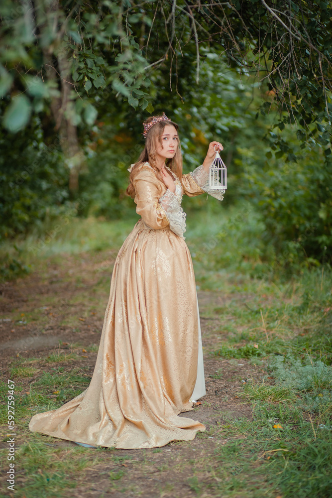 A beautiful young woman in a historical golden dress with a crown on her head holds a white cage in her hands. Princess in a medieval dress in the forest.
