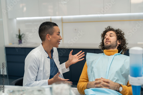 Photography Young woman dentist sitting in her dental care office next to male patient on dentist's chair, recommending and explaining dental procedures with smile, motivating patient before treatment