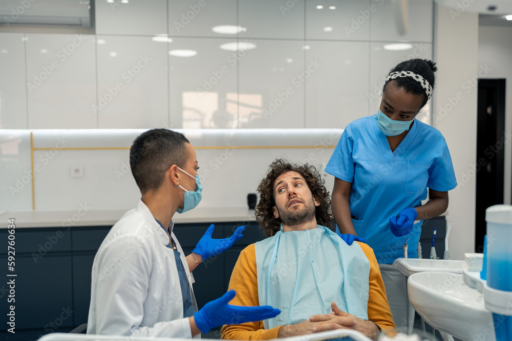 Obraz premium Anxious male patient on his first appointment with his dentist and her assistant dental nurse with surgical masks discussing future dental procedures and treatment for healthy gums and teeth.