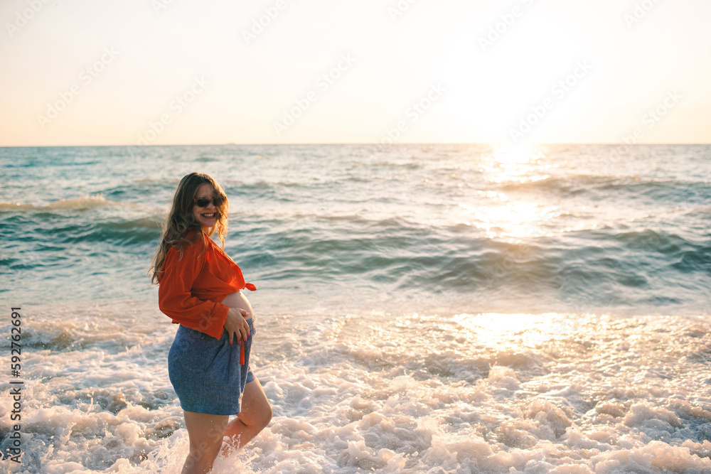 Pregnant happy woman walking at sea shore over sun light wearing casual prenatal clothes at beach. Motherhood. Summer vacation season. Pregnant happy woman walking at sea shore over sun light wearing casual prenatal clothes at beach. Motherhood. Summer vacation season.