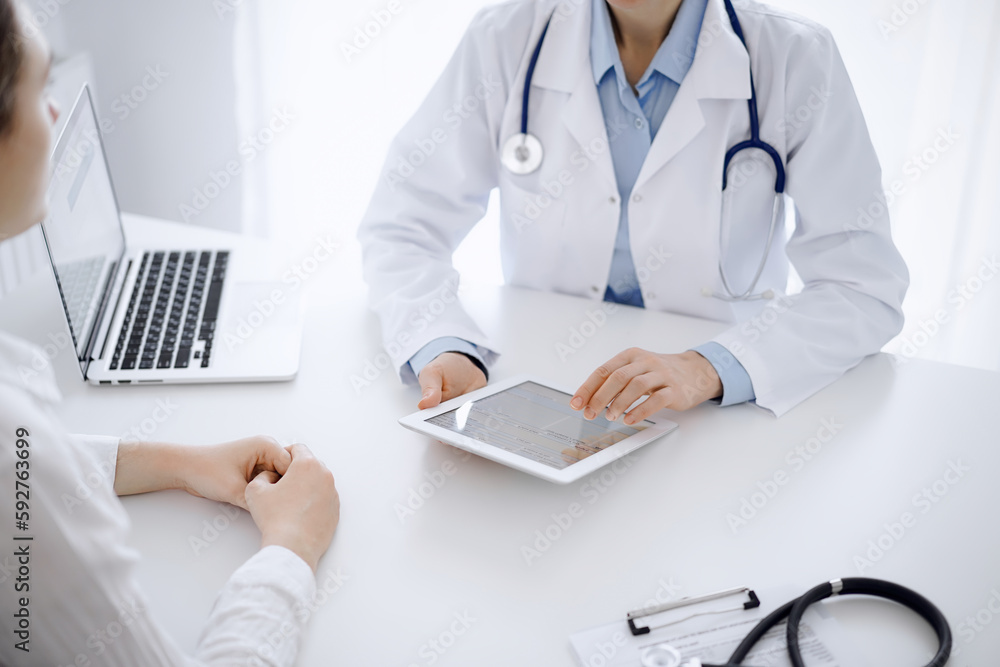 Doctor and patient sitting opposite each other at the desk in clinic. The focus is on female physician's hands pointing into tablet computer touchpad, close up. Medicine concept Doctor and patient sitting opposite each other at the desk in clinic. The focus is on female physician's hands pointing into tablet computer touchpad, close up. Medicine concept