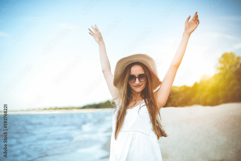 Happy smiling woman in free happiness bliss on ocean beach standing with a hat, sunglasses, and rasing hands. Portrait of a multicultural female model in white summer dress enjoying nature Happy smiling woman in free happiness bliss on ocean beach standing with a hat, sunglasses, and rasing hands. Portrait of a multicultural female model in white summer dress enjoying nature