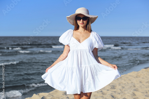 Happy smiling blonde woman is posing on the ocean beach with sunglasses and a hat
