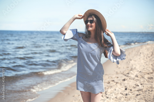 Happy young brunette woman standing on the ocean beach while smiling  and wearing fashion hat  sunglasses. The enjoying vacation concept