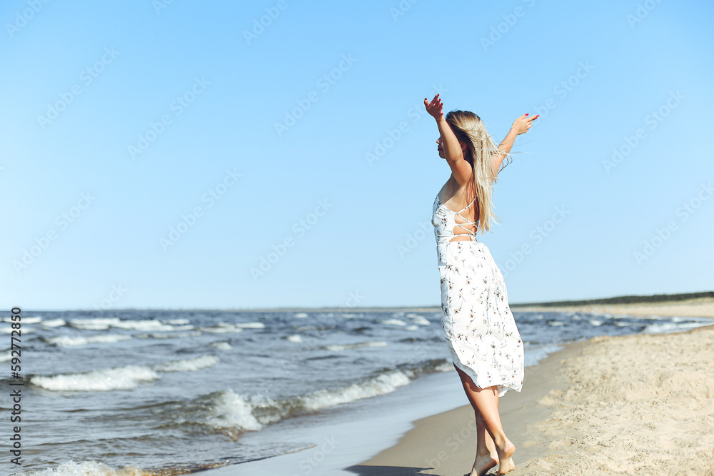 Happy blonde beautiful woman on the ocean beach standing in a white summer dress, open arms Happy blonde beautiful woman on the ocean beach standing in a white summer dress, open arms