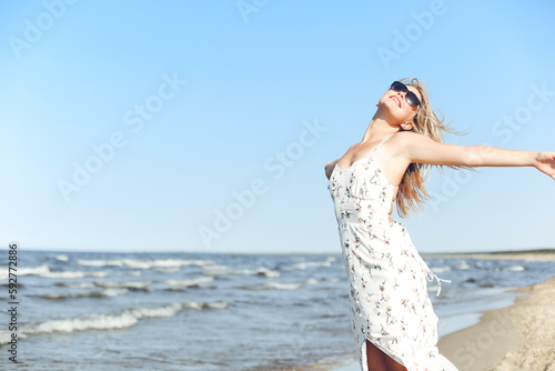 Happy blonde beautiful woman on the ocean beach standing in a white summer dress and sun glasses, open arms