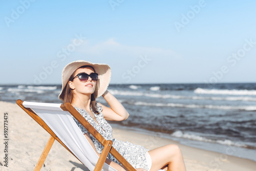 Happy brunette woman wearing sunglasses and hat relaxing on a wooden deck chair at the ocean beach