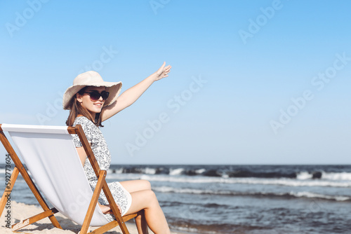 Happy brunette woman sitting on a wooden deck chair at the ocean beach while waving and greeting somebody with her hand © rogerphoto
