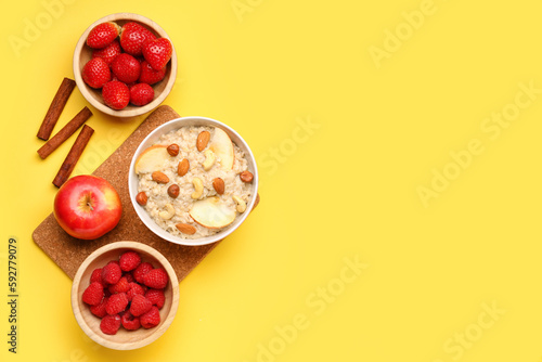 Bowl with tasty oatmeal, nuts, ripe berries, apple and cinnamon on yellow background