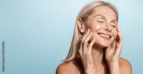 Gorgeous senior older woman with closed eyes touching her perfect skin. Beautiful portrait mid 50s aged woman advertising facial antiage lift products salon care tighten skin isolated on blue