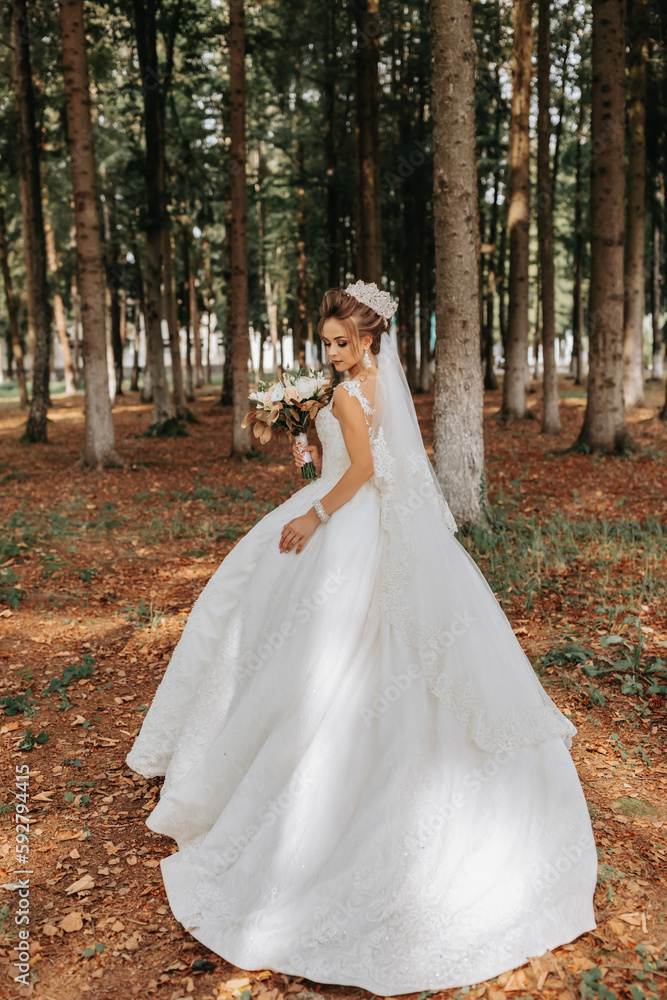 A beautiful young woman in a wedding dress between tall trees in the forest with a royal hairstyle and a chic tiara with a bouquet of flowers in her hands, a wedding in golden color