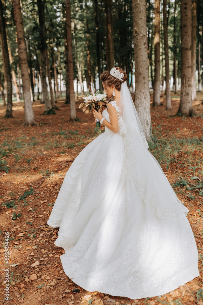 A beautiful young woman in a wedding dress between tall trees in the forest with a royal hairstyle and a chic tiara with a bouquet of flowers in her hands, a wedding in golden color A beautiful young woman in a wedding dress between tall trees in the forest with a royal hairstyle and a chic tiara with a bouquet of flowers in her hands, a wedding in golden color