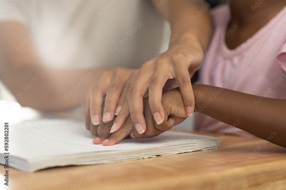 Biracial mother sitting at table helping her blind daughter to read braille Biracial mother sitting at table helping her blind daughter to read braille