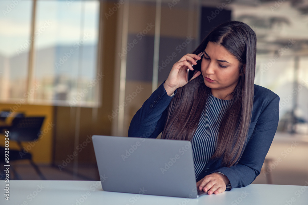 Serious businesswoman talking on mobile phone at office workplace, working at computer. Secretary answering telephone call. Serious businesswoman talking on mobile phone at office workplace, working at computer. Secretary answering telephone call.