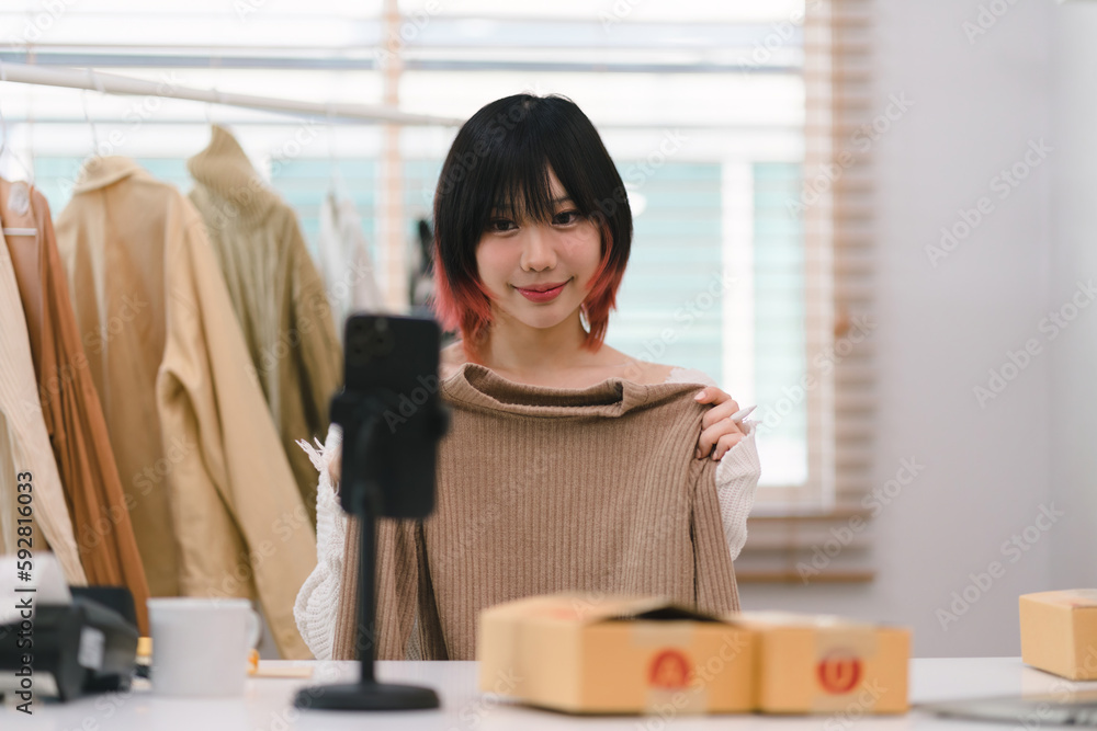 An Asian vlogger, blogger, small SME business owner who sells clothes online, is using her mobile phone for a live stream video call to present and sell her products in detail from her home office.