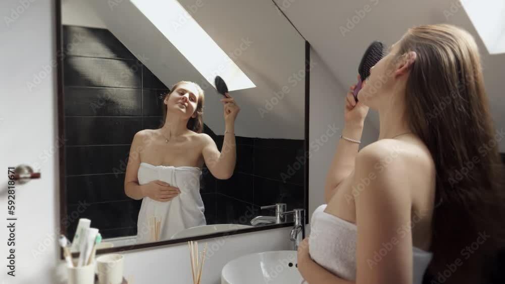 Woman standing in a bath, covered in a towel, and combing her long hair while looking at herself in the mirror. Concept of maintaining good hygiene and self-care rituals Woman standing in a bath, covered in a towel, and combing her long hair while looking at herself in the mirror. Concept of maintaining good hygiene and self-care rituals