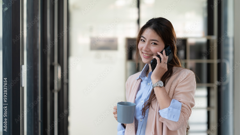 Asian businesswoman standing and holding a cup of coffee while talking smartphone in an office. Asian businesswoman standing and holding a cup of coffee while talking smartphone in an office.