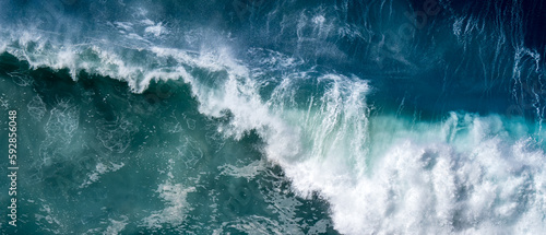 Sight from above as a big breaking wave crashes into the blue waters below with its power and strength visible through the white spray and foam, making it a awe moment captured in this panoramic view.