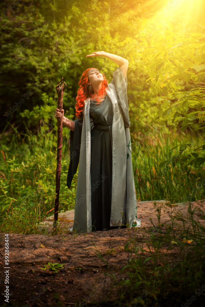 A young woman in a cloak, with a staff and a green dress in the form of a druid. Forest witch with bright red hair. Makeup with black pencil.