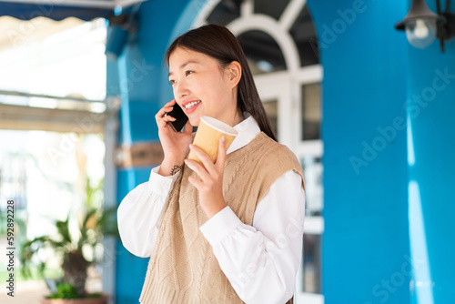 Young Chinese woman at outdoors using mobile phone and holding a coffee with happy expression photo