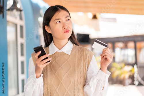 Young Chinese woman at outdoors buying with the mobile with a credit card while thinking photo