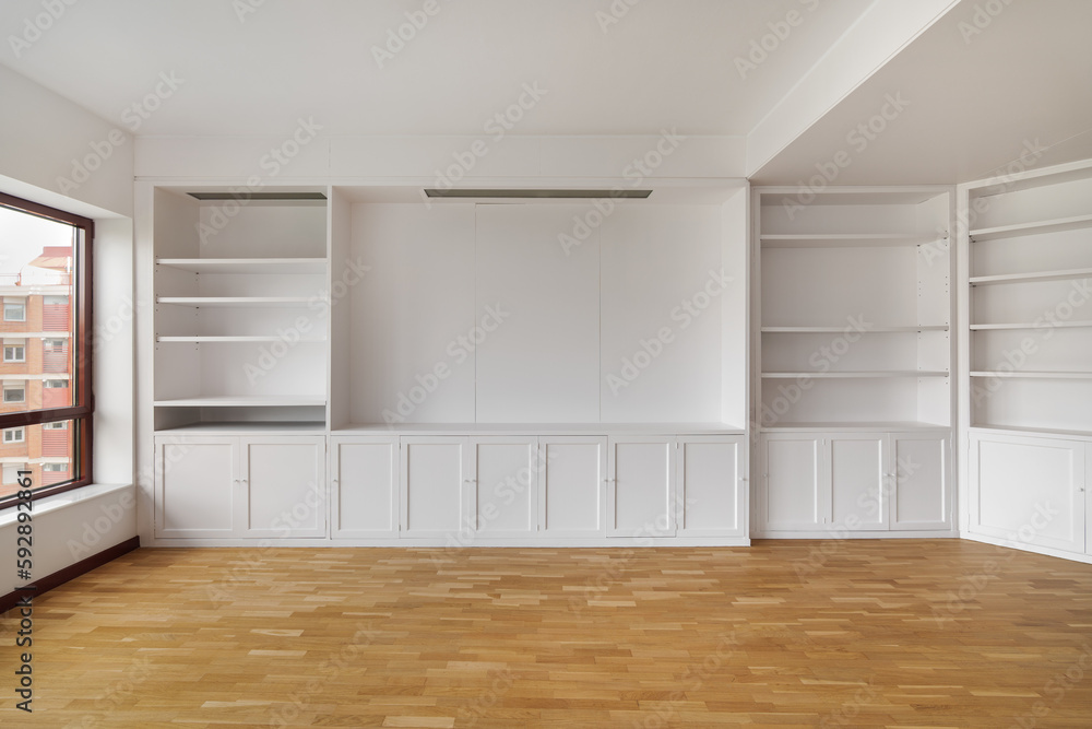 Fototapeta premium Empty built-in white cabinet on wooden laminate floor with big window in empty bright airy room in housewarming. Concept of housewarming and convenient storage