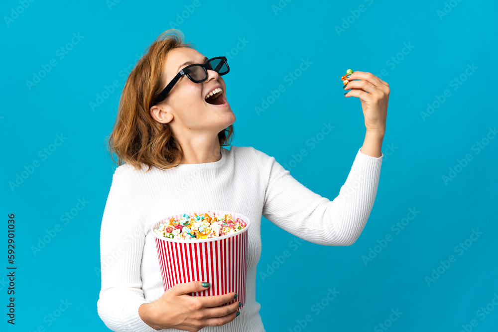 Young Georgian woman isolated on blue background with 3d glasses and holding a big bucket of popcorns