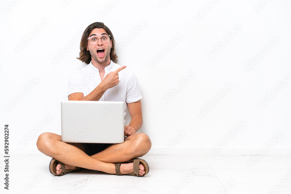 Young handsome man with a laptop sitting on the floor isolated on white background surprised and pointing side Young handsome man with a laptop sitting on the floor isolated on white background surprised and pointing side
