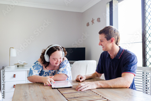 Teen wearing headphones talking with adult disability support worker in home office using tablet