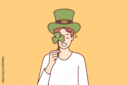 Happy man with St Patrick Day celebration accessories wearing green hat and covering eye with clover petal. Positive guy invites to traditional festival on eve of Irish holiday St Patrick Day