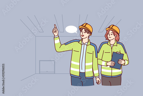 Two industrial room engineers discussing production process and how to repair equipment. Man and woman engineers in construction uniforms and safety helmets hold meetings in unfinished building 